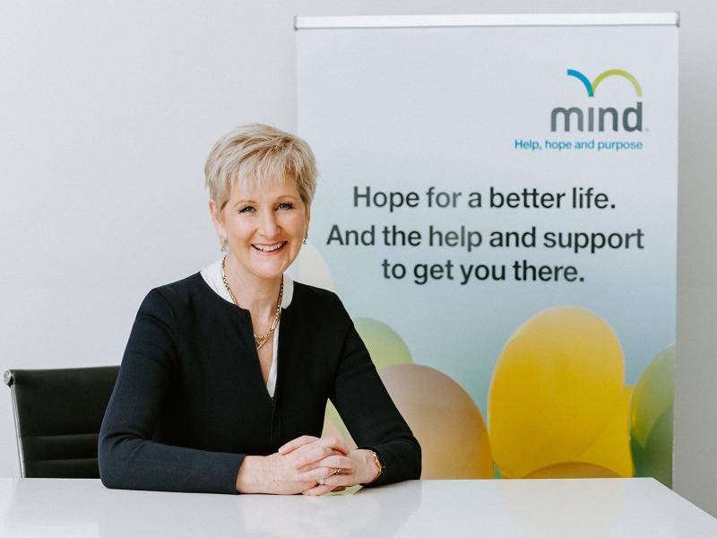 Gill Callister sitting at a desk. Wearing black with white collar. Smiling. Hands crossed.